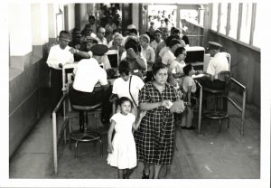 Immigration inspection at Southern land border port of entry 1947-1965. Source: United States Citizenship and Immigration Services History Office and Library. Black and white photograph; A women looks at the camera and so does a young girl to her right. There is a line of people behinf them.