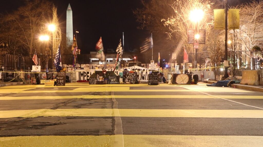 Night view of a section of fence blocking the road to the White House. A single line of caution tape stretches across the road in front of it. The fence is covered to the point of invisibility in anti-racist and anti-Trump protest banners and signs.